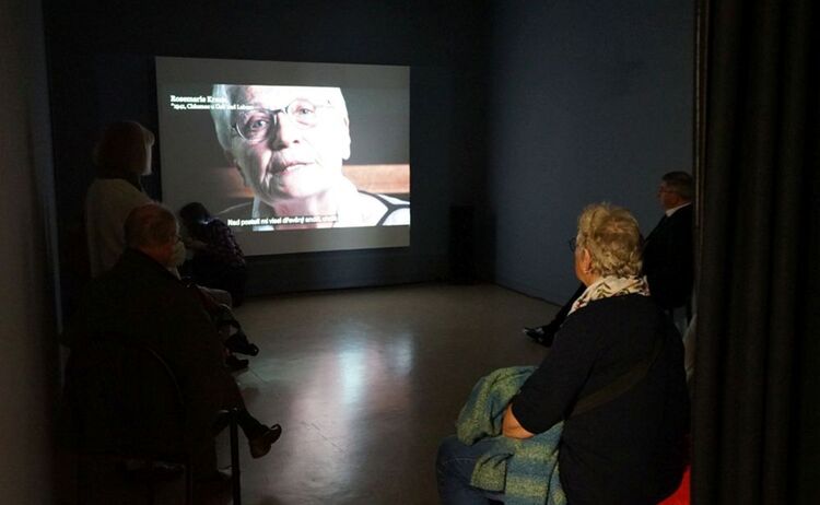 Besucherinnen und Besucher schauen das Zeitzeugenvideo in der Ausstellung: Zum Vergrößern auf Bild klicken