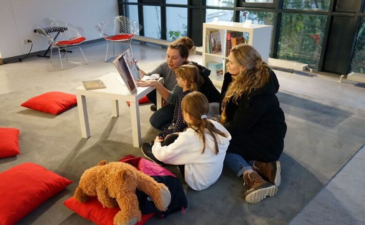 Zwei Besucherinnen mit zwei Mädchen in der Leseecke beim Lesen eines Buches.: Zum Vergrößern auf Bild klicken